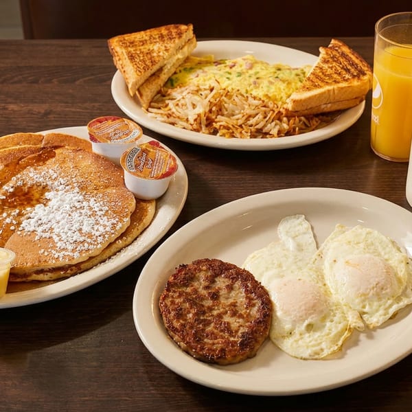 Hearty diner breakfast with pancakes, eggs, sausage, hash browns, toast, orange juice, and coffee on a wooden table.