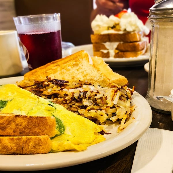 Diner breakfast plate with a cheesy omelet, toast, hash browns, coffee, juice, and layered dessert in the background.