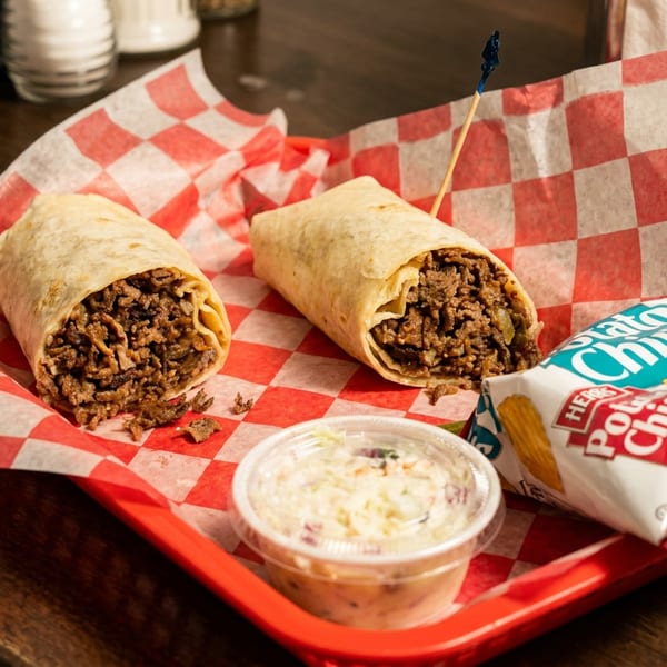Red tray holding two beef burritos, a cup of creamy dip, and a bag of potato chips on checkered paper.