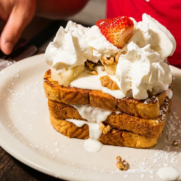Stacked French toast topped with whipped cream, strawberries, walnuts, and powdered sugar on a plate.