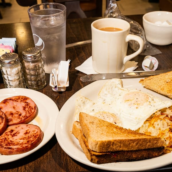 Diner breakfast featuring eggs, toast, hash browns, grilled ham slices, coffee, and condiments on a wooden table.