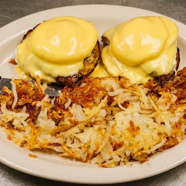 Plate of eggs Benedict with golden hollandaise sauce, served alongside crispy shredded hash browns on a skillet.