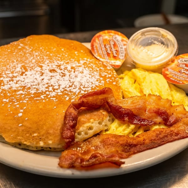 Breakfast plate with powdered-sugar pancakes, scrambled eggs, crispy bacon, and individual Smucker’s syrup cups.